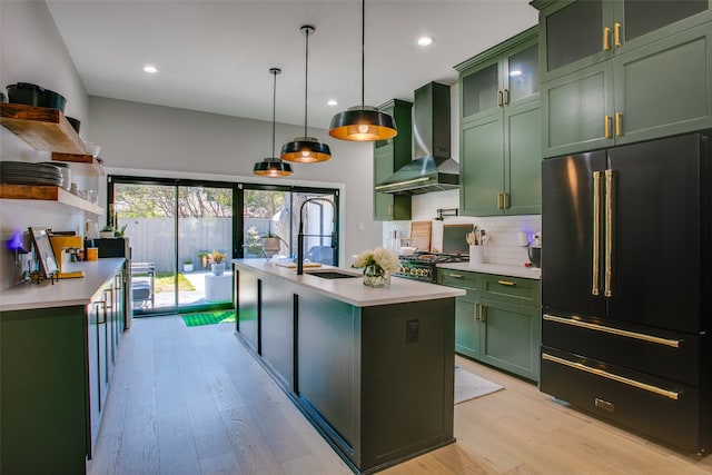 kitchen with high end refrigerator, wall chimney range hood, a center island with sink, light hardwood / wood-style flooring, and green cabinets