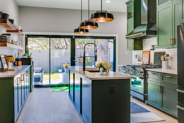 kitchen featuring pendant lighting, wall chimney range hood, stainless steel stove, a kitchen island with sink, and green cabinetry