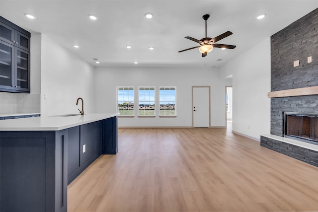 kitchen with sink, backsplash, an island with sink, a stone fireplace, and light wood-type flooring