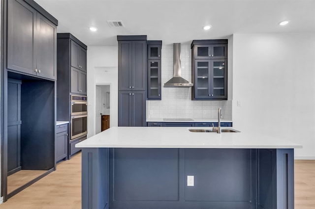 kitchen with black electric cooktop, a kitchen island with sink, wall chimney exhaust hood, and light wood-type flooring