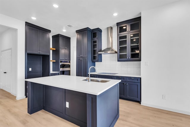 kitchen with sink, a center island with sink, black electric cooktop, wall chimney range hood, and backsplash