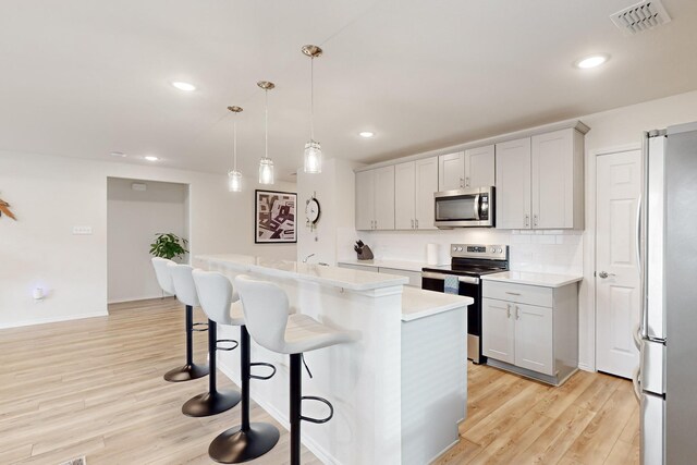 kitchen with light wood finished floors, an island with sink, decorative light fixtures, stainless steel appliances, and light countertops