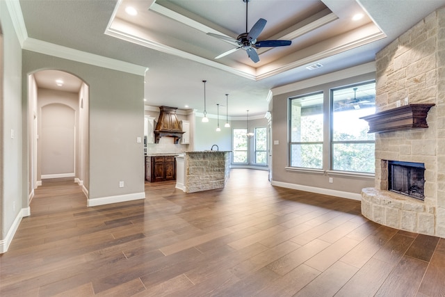 unfurnished living room with wood-type flooring, a stone fireplace, ceiling fan, and a tray ceiling