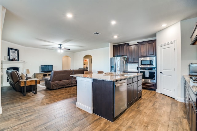 kitchen with wood-type flooring, appliances with stainless steel finishes, a center island with sink, and dark brown cabinetry