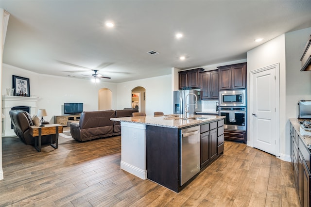 kitchen with ceiling fan, wood-type flooring, a center island with sink, dark brown cabinets, and appliances with stainless steel finishes