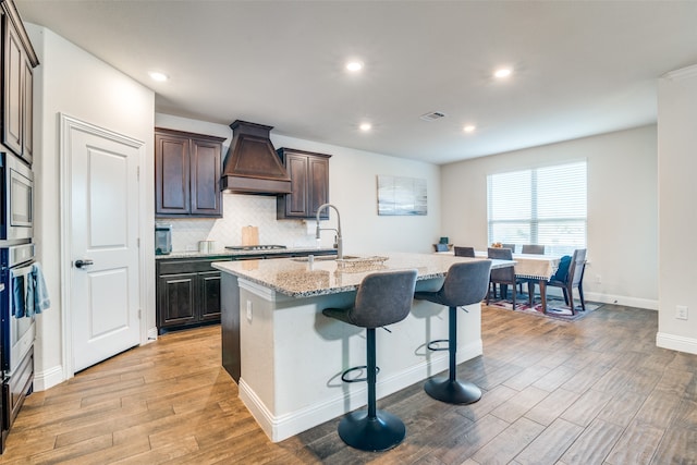 kitchen with a kitchen breakfast bar, light wood-type flooring, custom exhaust hood, stainless steel appliances, and a kitchen island with sink