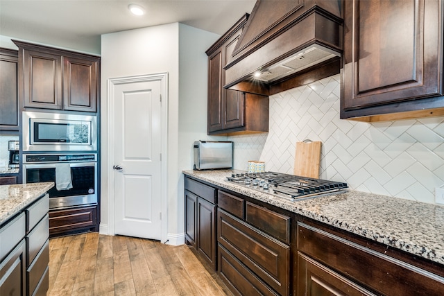 kitchen with dark brown cabinetry, light stone counters, premium range hood, appliances with stainless steel finishes, and light wood-type flooring