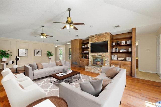 living room featuring built in shelves, lofted ceiling, ceiling fan, a fireplace, and light hardwood / wood-style floors