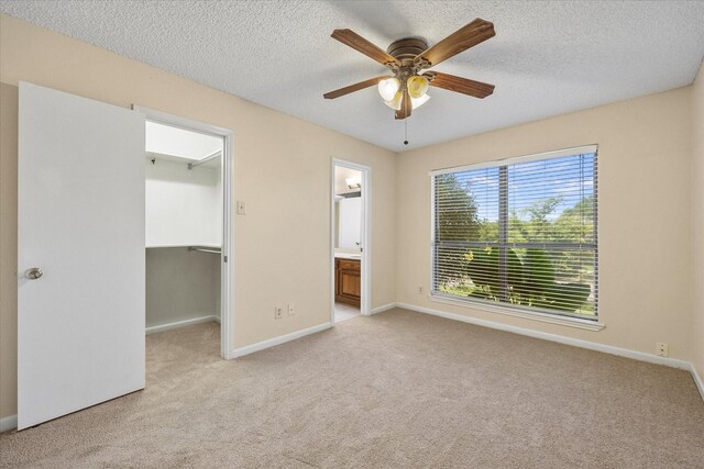 unfurnished bedroom featuring ceiling fan, ensuite bathroom, a textured ceiling, a walk in closet, and light carpet