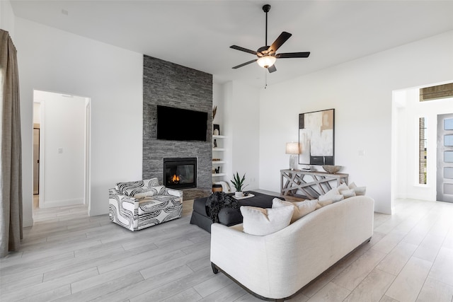 living room with ceiling fan, a stone fireplace, built in shelves, and light hardwood / wood-style flooring