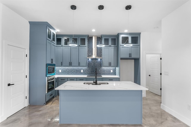 kitchen with light stone counters, wall chimney exhaust hood, stainless steel appliances, a kitchen island with sink, and hanging light fixtures