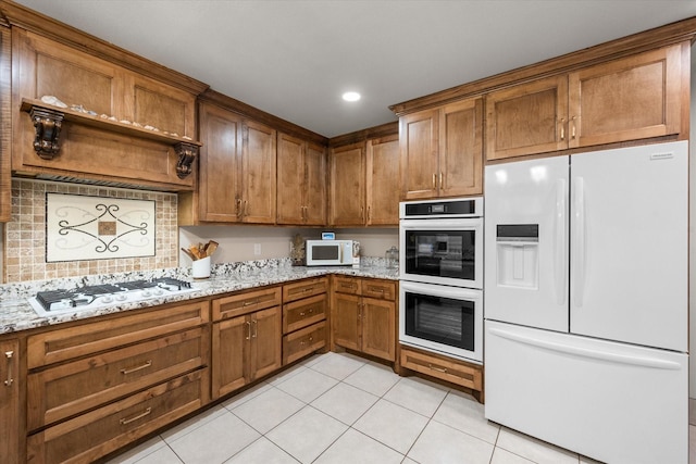 kitchen with brown cabinets, light tile patterned flooring, light stone counters, and white appliances
