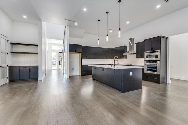 kitchen with sink, decorative light fixtures, a center island with sink, double oven, and wall chimney range hood