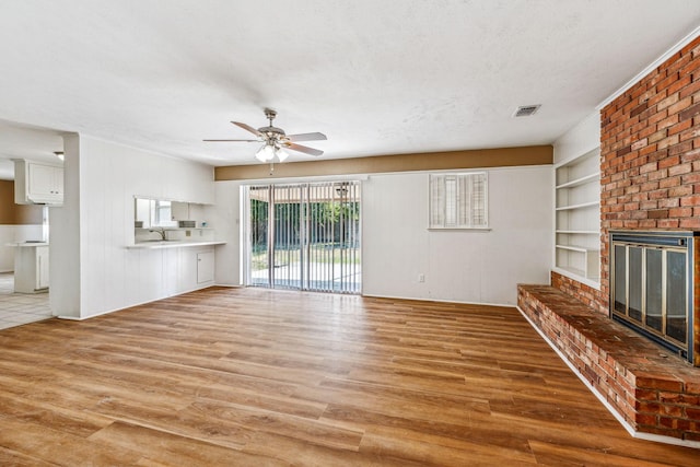 unfurnished living room featuring a brick fireplace, light hardwood / wood-style flooring, ceiling fan, built in shelves, and a textured ceiling