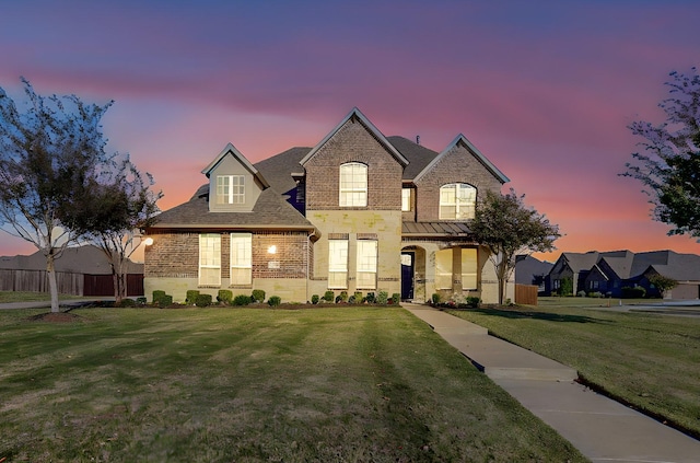 french country inspired facade with brick siding, fence, a front lawn, and roof with shingles