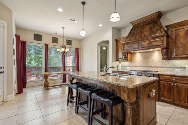 kitchen with sink, hanging light fixtures, light stone counters, premium range hood, and a center island with sink