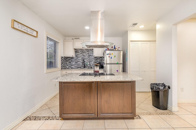 kitchen featuring light stone counters, island range hood, light tile patterned floors, stainless steel fridge, and an island with sink