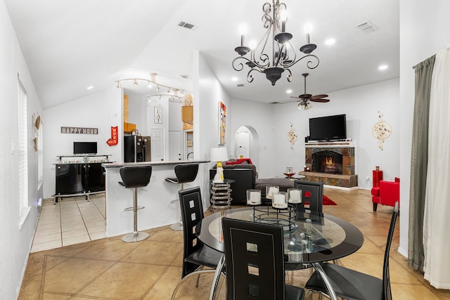 tiled dining space with ceiling fan with notable chandelier, vaulted ceiling, and a stone fireplace