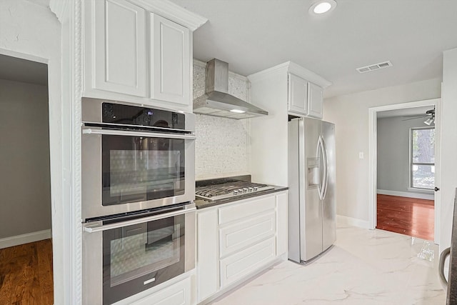 kitchen featuring wall chimney range hood, light hardwood / wood-style floors, decorative backsplash, white cabinets, and appliances with stainless steel finishes