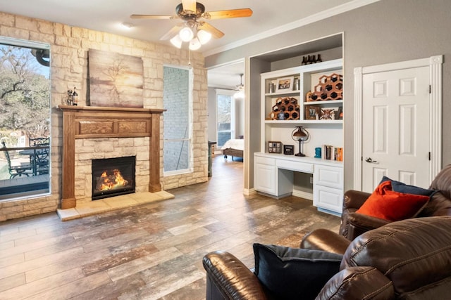 living room with built in desk, a fireplace, wood-type flooring, ornamental molding, and ceiling fan