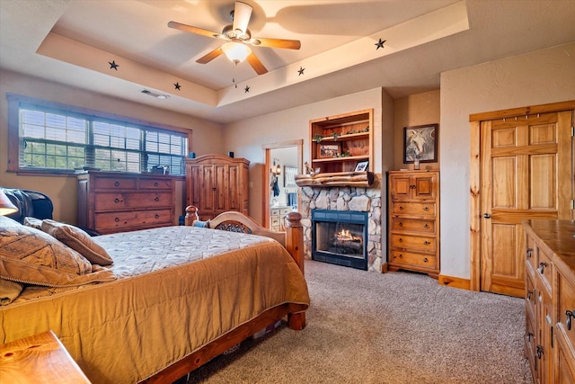 carpeted bedroom with a tray ceiling, a stone fireplace, and ceiling fan