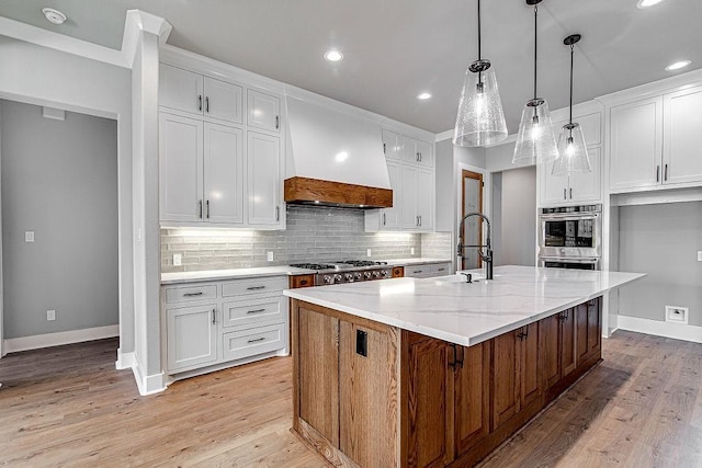 kitchen featuring premium range hood, a kitchen island with sink, light hardwood / wood-style flooring, light stone countertops, and white cabinetry