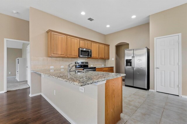 kitchen with light stone countertops, light hardwood / wood-style flooring, kitchen peninsula, decorative backsplash, and appliances with stainless steel finishes