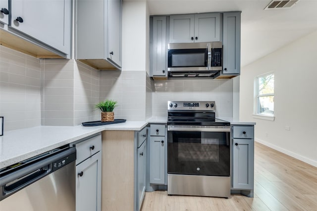 kitchen with gray cabinetry, light stone countertops, tasteful backsplash, appliances with stainless steel finishes, and light wood-type flooring