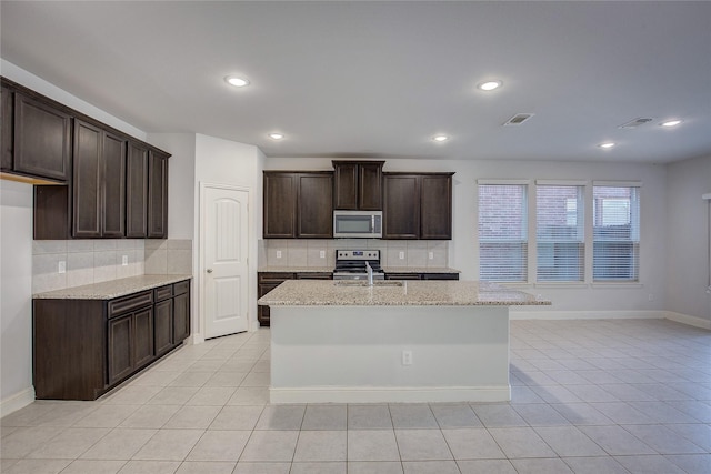 kitchen featuring light stone counters, dark brown cabinetry, stainless steel appliances, and a center island with sink