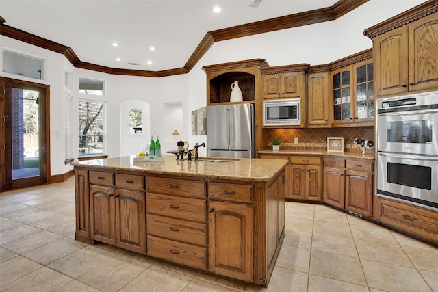 kitchen with light stone countertops, appliances with stainless steel finishes, ornamental molding, sink, and an island with sink