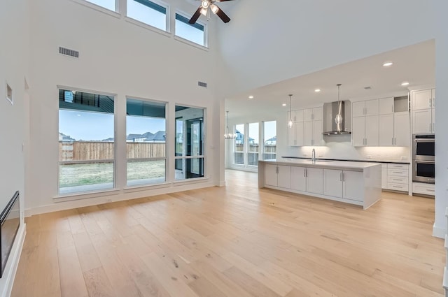 kitchen with an island with sink, white cabinets, decorative light fixtures, wall chimney exhaust hood, and light wood-type flooring