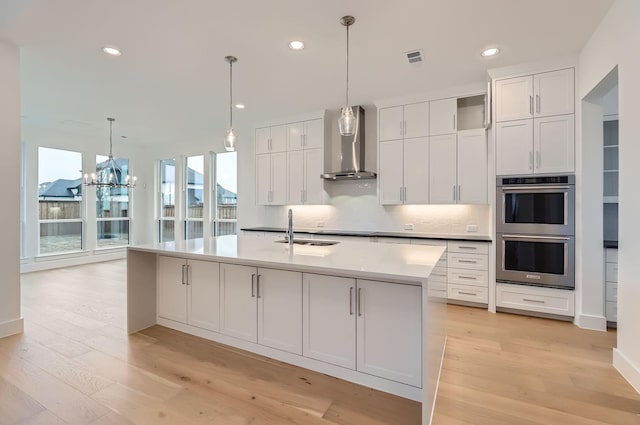 kitchen featuring wall chimney exhaust hood, a center island with sink, pendant lighting, stainless steel double oven, and white cabinets