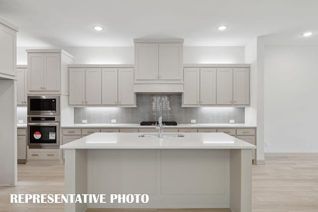 kitchen with backsplash, stainless steel appliances, sink, light hardwood / wood-style floors, and an island with sink