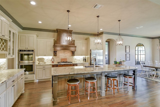 kitchen with stainless steel appliances, dark hardwood / wood-style flooring, premium range hood, a large island with sink, and decorative light fixtures