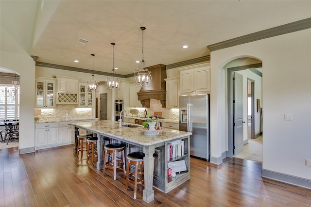 kitchen with pendant lighting, premium range hood, a kitchen island with sink, hardwood / wood-style flooring, and appliances with stainless steel finishes