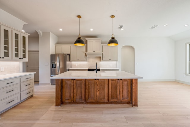 kitchen with sink, a kitchen island with sink, stainless steel refrigerator with ice dispenser, light stone countertops, and decorative light fixtures