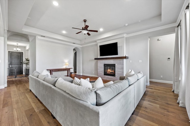 living room featuring ceiling fan with notable chandelier, a tray ceiling, a stone fireplace, and hardwood / wood-style flooring