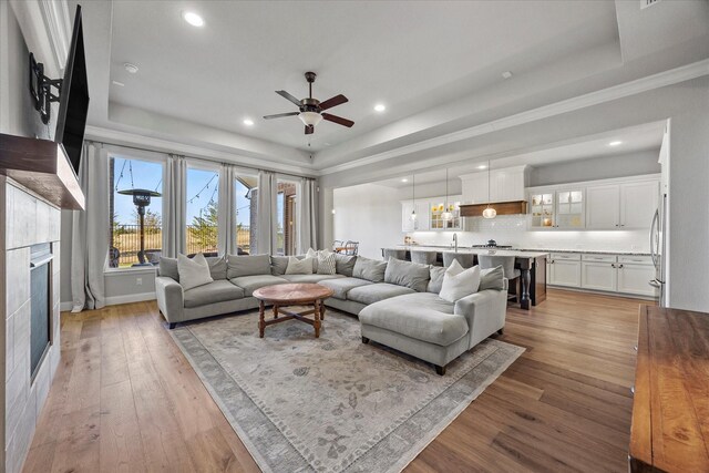 living room featuring ceiling fan, sink, a raised ceiling, light hardwood / wood-style floors, and a tiled fireplace