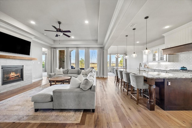 living room featuring light hardwood / wood-style flooring, a tray ceiling, a tiled fireplace, ceiling fan with notable chandelier, and ornamental molding