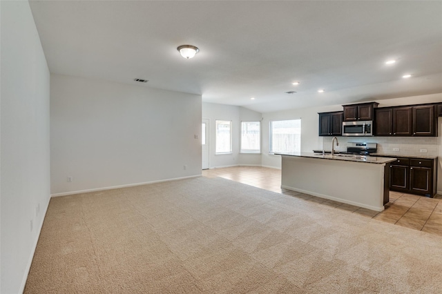 kitchen featuring dark brown cabinets, light colored carpet, an island with sink, and appliances with stainless steel finishes