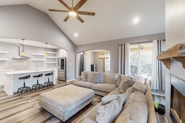 living room featuring built in shelves, sink, lofted ceiling, ceiling fan with notable chandelier, and light wood-type flooring