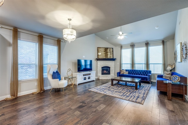 living area featuring baseboards, dark wood-style flooring, a stone fireplace, vaulted ceiling, and ceiling fan with notable chandelier