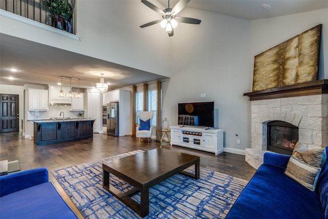 living area with baseboards, a stone fireplace, ceiling fan with notable chandelier, a towering ceiling, and dark wood-style floors