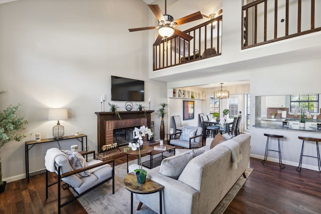 living room with ceiling fan with notable chandelier, dark wood-type flooring, a high ceiling, and a brick fireplace