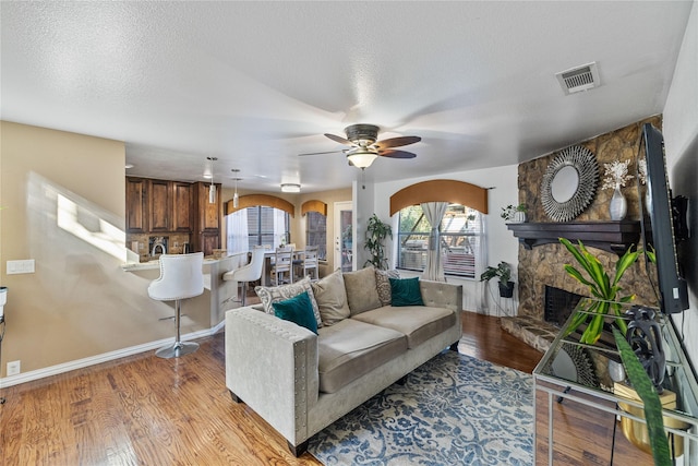 living room featuring ceiling fan, a stone fireplace, a textured ceiling, and light hardwood / wood-style flooring