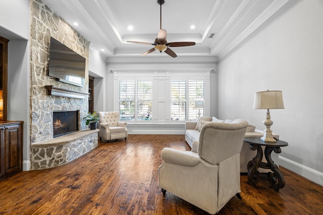 living room with ceiling fan, a stone fireplace, dark hardwood / wood-style flooring, a tray ceiling, and ornamental molding