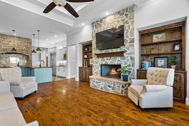 living room with a stone fireplace, sink, crown molding, ceiling fan, and dark hardwood / wood-style flooring