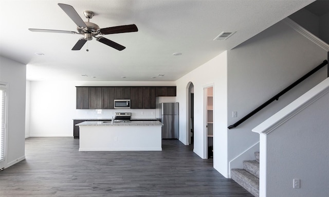 kitchen with ceiling fan, dark wood-type flooring, an island with sink, dark brown cabinets, and appliances with stainless steel finishes