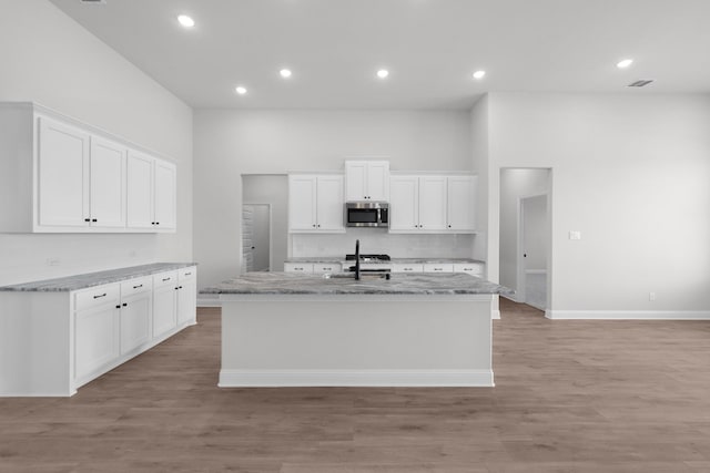 kitchen with a center island with sink, a sink, white cabinets, stainless steel microwave, and light wood-type flooring