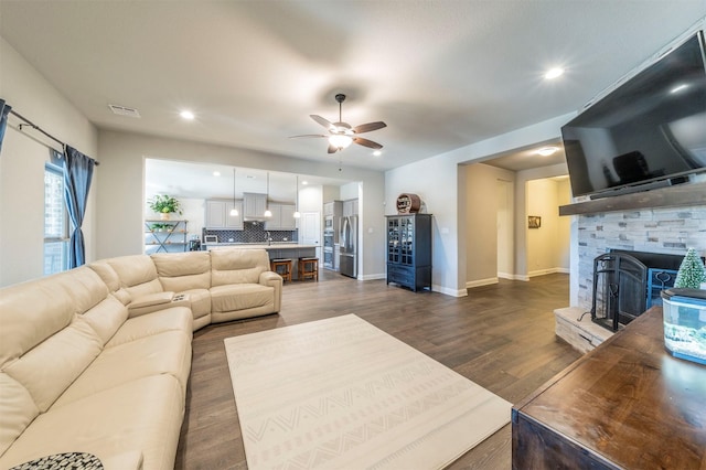 living room with a stone fireplace, dark wood-type flooring, and ceiling fan